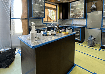 A kitchen under renovation with furniture covered in plastic, various tools and materials on the island, and protective tape lining the edges and floor.