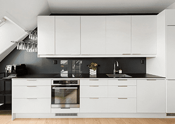 Modern kitchen with white cabinets, black countertop, stainless steel sink and appliances, including an oven and built-in microwave. Wine glasses hang under slanted ceiling. Light wood floor.