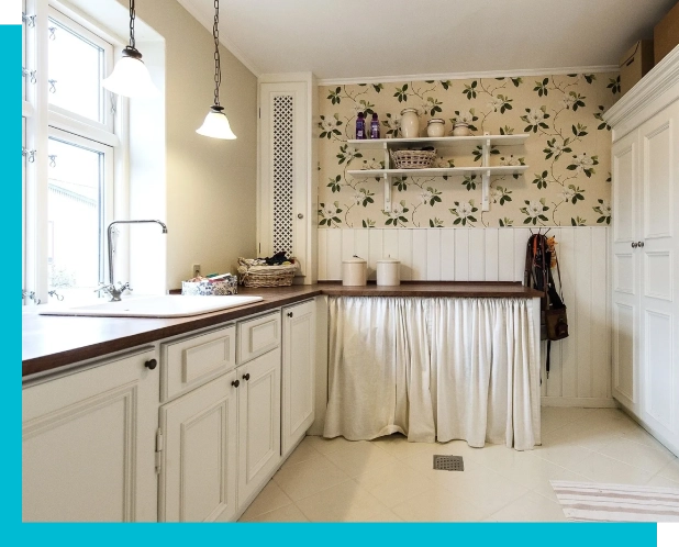 A kitchen with white cabinets, wooden countertops, a sink by the window, decorative wallpaper, open shelves with items, and storage hidden by white curtains below the counter. Two pendant lights hang above.