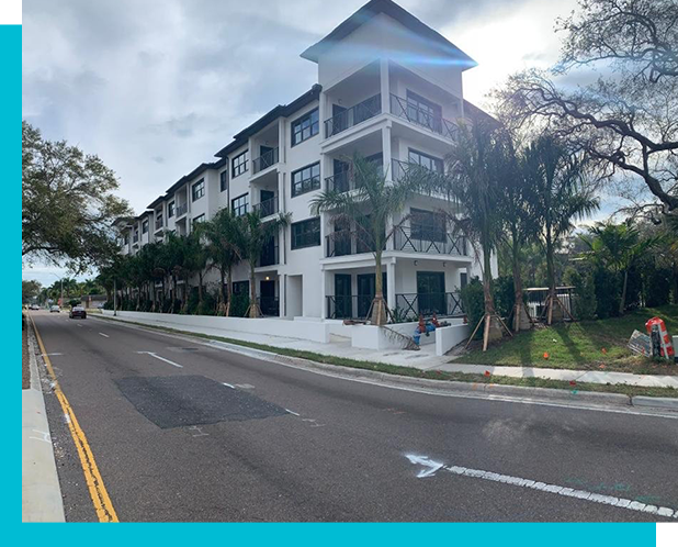 A four-story modern apartment building with white walls and dark trim stands along a quiet street, with palm trees and light landscaping in front.