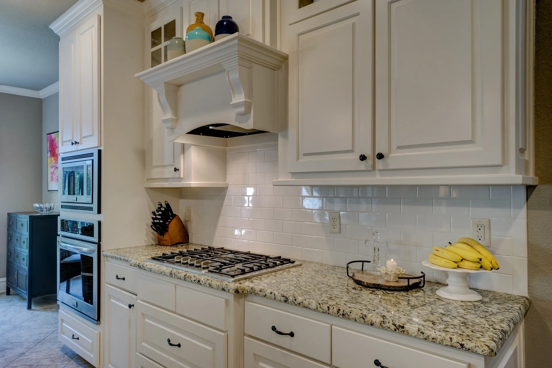 A modern kitchen featuring white cabinets, a granite countertop, a gas stovetop, and a built-in oven. A knife block and a bowl of bananas are on the counter. White subway tile backsplash adds a clean look.