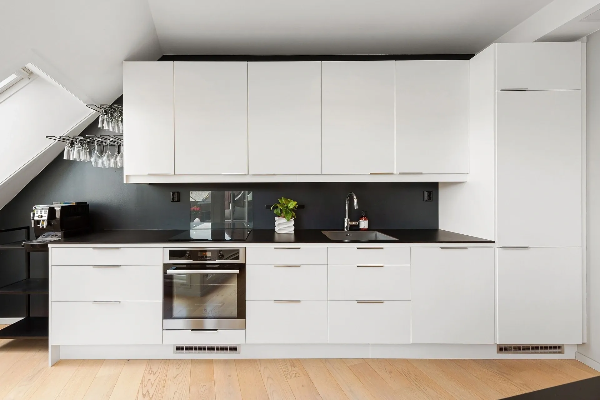 Modern kitchen with white cabinets, black countertops, built-in oven, small sink, and stainless-steel fixtures. A coffee machine, wine glasses, and plant are visible on the countertop.