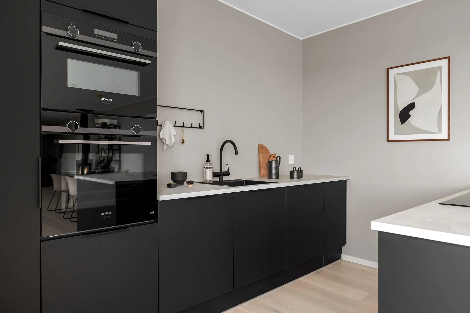 A modern kitchen with black cabinets, a built-in oven, a sink, and a minimalist faucet. White countertop contrasts with the cabinets, and a framed abstract artwork hangs on the right wall.