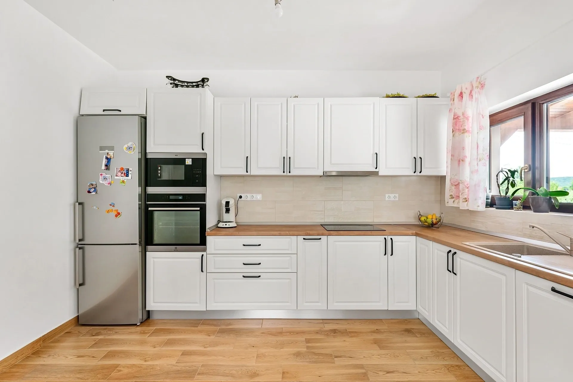 A modern kitchen with white cabinets, a double oven, a refrigerator with magnets, and a wooden floor. There is a window with pink curtains and a countertop with a fruit bowl and kitchen items.