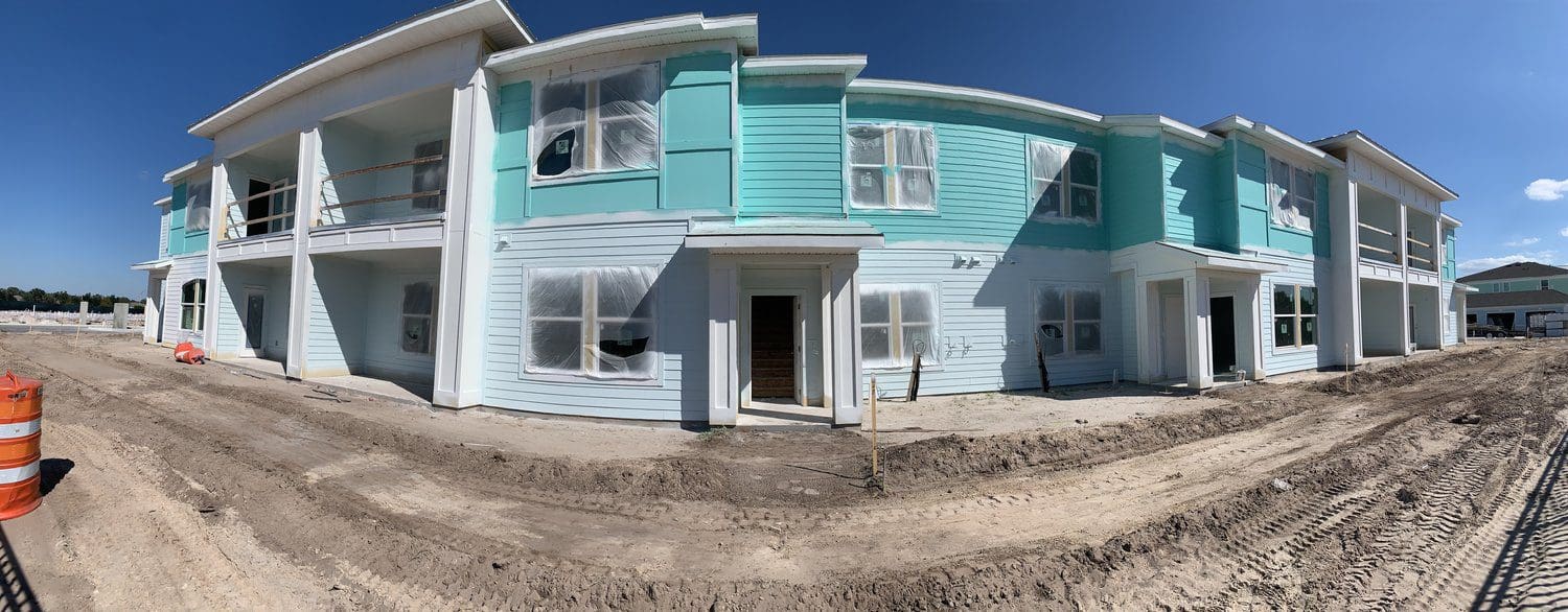 A partially constructed two-story building with turquoise and white exterior, surrounded by dirt. Windows are covered, and orange construction barrels are present. Clear blue sky in the background.